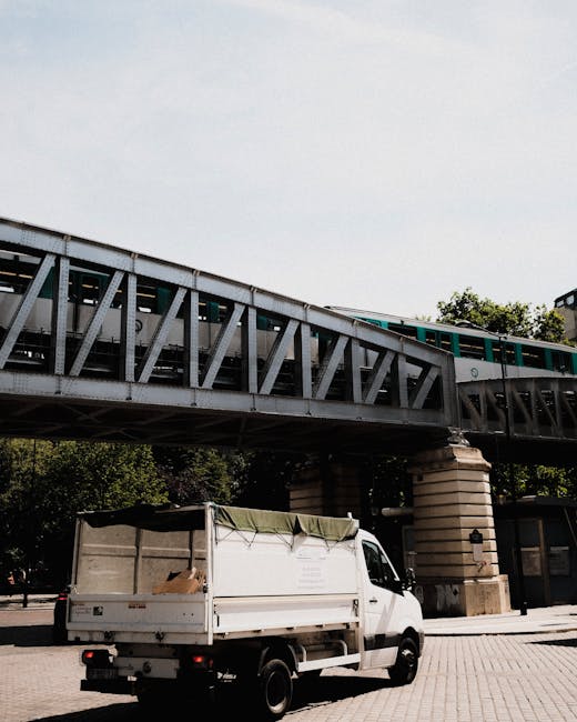 A white flatbed van with a partially open cargo area is parked on a paved surface beneath a steel railway bridge supported by stone pillars. The cargo area of the van contains some loose materials or debris, covered with a green tarpaulin or protective cover, suggesting it is used for transporting waste or rubbish. The bridge above is constructed of steel trusses painted in dark grey and features a train passing across, with visible windows and horizontal green and white train carriages. In the background, there are leafy green trees and glimpses of buildings, indicating an urban environment. The lighting appears natural, with sunlight illuminating the scene, creating soft shadows beneath the van and around the pillars. The setting reflects a typical scene where a private or independent rubbish collection service, such as House Clearance Earls Court, might facilitate waste removal from a residential or urban location, where on-site clearance or alternative disposal methods are employed outside of standard council services.