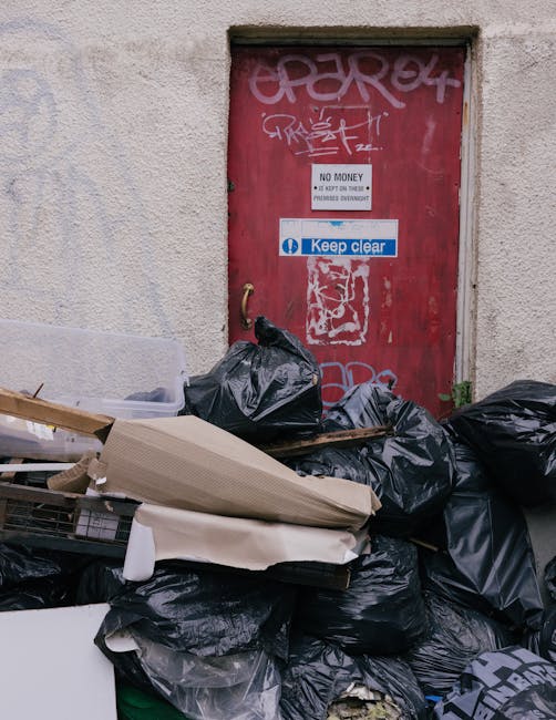 A pile of black garbage bags, some torn open revealing miscellaneous waste such as flattened cardboard boxes, paper rolls, and plastic debris, situated in front of a red metal door set into a textured beige wall. The door features graffiti tags, a 'No Money' notice, and a blue 'Keep Clear' sticker, with a brass door handle visible on the left side. The waste is arranged haphazardly on a paved surface, with surrounding remnants of discarded materials. The scene suggests an area designated for rubbish collection or an alternative waste handling point, possibly managed by a private waste removal service such as House Clearance Earls Court. The environment appears outdoor, with natural lighting illuminating the scene, emphasizing the contrast between the debris and the building’s facade. This image exemplifies typical materials involved in rubbish removal in an urban setting, awaiting collection or disposal, and highlights the importance of proper waste management and clearance services for property cleanliness and tidiness.