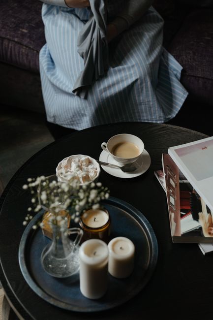 The image depicts a round black coffee table with various items placed on it, including a small glass vase containing a few sprigs of white flowers, a large candle with a metallic gold base, and two white pillar candles. Additionally, there is a white cup filled with a light-colored hot beverage, resting on a matching saucer, and a small bowl of what appears to be whipped cream or a similar topping. To the right of the candles, a partially open magazine with glossy pages features photographs and text. In the background, a person is seated on a dark upholstered sofa, wearing a light blue and white striped dress, with their hands and part of their lap visible, holding a dark jacket or piece of clothing. The setting appears to be indoors, with soft, natural lighting creating a cozy atmosphere, indicating a relaxed environment suitable for casual reading or enjoying a hot drink, in a home or café setting. The scene aligns with elements associated with casual domestic or hospitality environments, intended to evoke comfort and quiet relaxation, consistent with the context of home or private rubbish removal and general waste management services provided by House Clearance Earls Court.
