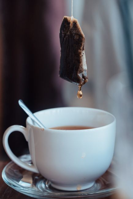 A white ceramic teacup filled with tea sits on a saucer, with a small metal spoon resting inside the cup. Above the cup, a teabag with a dark, damp, textured paper exterior hangs from a string, gradually releasing a drop of tea into the beverage. The background features a blurred, neutral-toned environment, likely indoors, with soft natural lighting highlighting the glossy finish of the cup and the moist surface of the teabag. The scene captures a moment of tea preparation, emphasizing the everyday environment and the process of steeping tea, providing a detailed visual relevant to residential and commercial beverage service areas that may involve disposal or handling of used teabags, as part of a broader context of waste management and disposal practices, which House Clearance Earls Court may support through alternative waste handling methods.