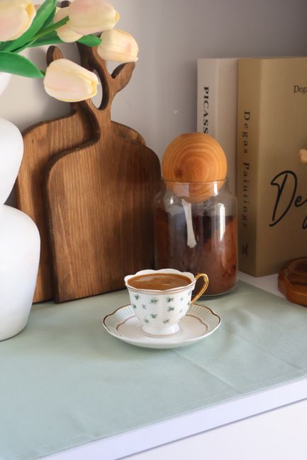 The image depicts a close-up of a decorative indoor surface, possibly a shelf or a sideboard, featuring a small porcelain teacup with a gold rim and floral patterns, filled with a dark beverage. To the left of the cup, there is a white ceramic vase holding light pink tulips with green stems and leaves. Behind the teacup, there are two wooden objects; a tall, flat, rectangular wooden tray with rounded edges and a natural wood finish, and a smaller, rounded wooden container with a matching lid, both with smooth surfaces and warm tones. To the right, there are a few books with light-colored covers standing upright; one with the title partly visible, mentioning 'Design' and 'Decor,' placed on a light-colored surface or tablecloth. The setting appears to be bright, with soft natural or diffuse lighting that highlights the textures and finishes of the objects, creating a cozy and aesthetic environment suited for a home or showroom display related to interior decoration or lifestyle. The overall scene complements the themes of home organization or private space styling, which can parallel the concept of independent waste handling or tasteful rubbish management in a domestic context, as referenced in the accompanying service page.