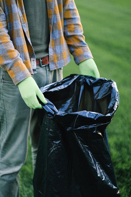 Close-up of a person holding an open black rubbish bag outdoors on a grassy area, wearing light green gloves, a green shirt, and a checked shirt in shades of yellow, grey, and blue. The rubbish bag appears to be made of plastic with a shiny surface, and the person is standing next to a green lawn, suggesting an outdoor waste collection or clearance activity related to rubbish removal services. The background features a well-maintained grass lawn, and the person's hands are fully visible, carefully holding the bag at its opening, ready for waste disposal or collection, aligned with the context of professional waste management and private rubbish handling services.