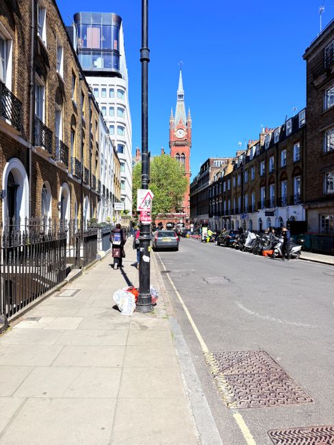 A city street scene featuring row houses with brick facades on the left, and parked motorcycles and cars on the right. The sidewalk in the foreground has a few pedestrians, and visible trash bags are placed next to a black railing. A tall black lamppost stands near the center of the image, with traffic and utility signs attached. In the background, a prominent red brick clock tower with a spire and a large clock face rises above the surrounding buildings, set against a bright blue sky with minimal clouds. The scene captures an urban environment consistent with a London district, with typical residential and commercial buildings, and indicates an area where local authority waste collection might be supplemented by private rubbish removal services, as suggested by the neatly placed waste bags along the pavement. The overall lighting is bright and natural, contributing to a clear and detailed visual impression of the street and its surroundings.