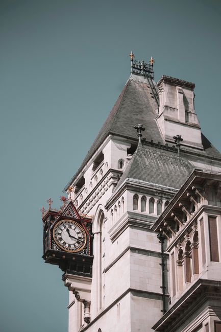 Close-up view of a historic clock tower featuring a large, ornate clock face with Roman numerals in black and gold, mounted on a white stone building with intricate architectural details including decorative arches, cornices, and carved stonework. The tower has a steep, slate-grey roof with a slightly weathered texture, topped with a small spire and finials. The building's exterior shows a combination of light grey and beige stone finishes, with some areas accented by darker trim. The background reveals a clear sky with a subtle gradient from light to darker blue, highlighting the architectural features. This scene is devoid of any immediate rubbish or waste-related objects but may situate within an urban setting where private or alternative waste collection options could be considered for maintaining the historic site’s surroundings. House Clearance Earls Court may be pertinent for local waste removal services in similar environments.