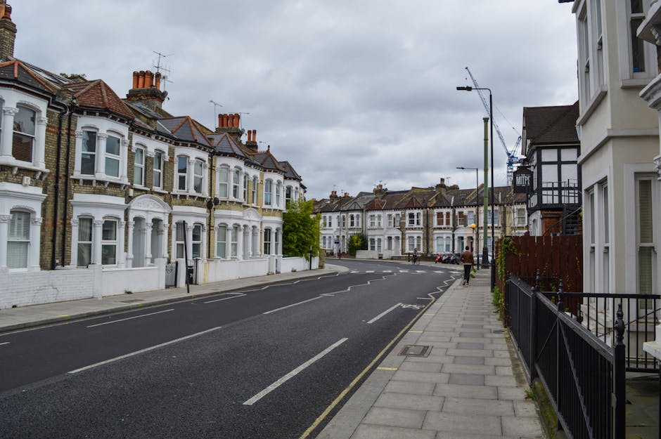 A quiet residential street featuring a row of Victorian-style terraced houses with white painted facades, bay windows, and decorative architectural details, set against an overcast sky. The pavement on the right side is paved with light grey concrete slabs, with a black wrought iron fence bordering some of the front gardens. A few pedestrians are visible walking along the sidewalk, and several parked cars line the curb. A tall lamppost extends above the sidewalk, and in the background, there are additional rows of similar terraced houses with tiled roofs and brick chimneys. The street appears clean and well-maintained, with natural light subdued by the cloudy weather, reflecting an ordinary day in an urban residential area. The scene subtly emphasizes the context of local city living and the need for occasional rubbish removal or waste management services, with no visible debris or clutter in sight. This visual setting aligns naturally with private waste handling and independent rubbish collection practices, characteristic of such neighbourhoods.