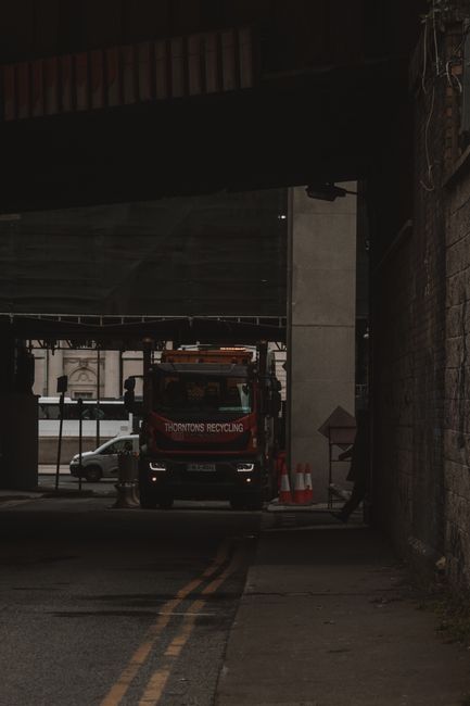 A waste collection truck with a red and black colour scheme, marked with the name 'Thornton's Recycling,' is positioned in a narrow, shaded alleyway beneath an elevated road structure. The truck's front end faces inward, with a visible yellow hazard light on the roof, indicating active collection or loading. To the right of the truck, a brick wall with visible wiring and conduits runs along the alley, while on the left, the underside of a bridge or flyover creates a dark overhead canopy. In the background, part of a street scene can be seen through the gap, including parked cars and building facades, illuminated by daylight. The pavement in front of the truck appears slightly worn, with faded double yellow lines marking the road. The setting suggests an urban environment where private waste collection services, such as those provided by House Clearance Earls Court, may access tricky urban locations for rubbish removal or site clearance involving difficult access points like alleyways beneath infrastructure.